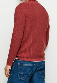 Roter gestrickter Pullover mit strukturiertem Muster, rundem Ausschnitt und gerippten Bündchen. Über blauen Jeans getragen, Rückansicht gezeigt.