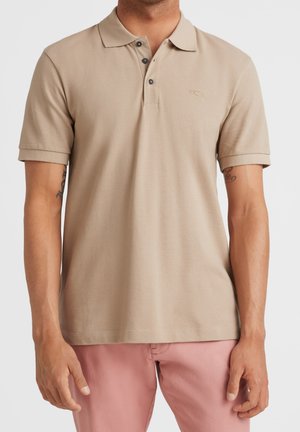 Mężczyzna w beżowej koszulce polo z krótkim rękawem, z trzema czarnymi guzikami, ubrany w jasnoróżowe spodnie, stoi z ramionami rozluźnionymi.