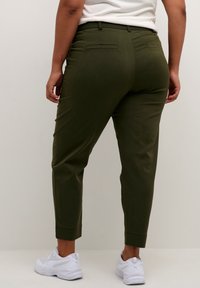 Pantalon vert olive, coupe ajustée, avec passants pour ceinture et deux poches arrière, assorti à des chaussures de sport blanches. Texture lisse.