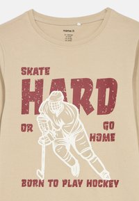 Μπεζ μπλούζα φτιαγμένη από βαμβάκι με στρογγυλή λαιμόκοψη, που διαθέτει γραφικό ενός παίκτη χόκεϊ και έντονο κόκκινο κείμενο: "ΚΑΝΕ ΚΑΛΟ ΣΚΕΪΤ Ή ΠΗΓΑΙΝΕ ΣΠΙΤΙ."