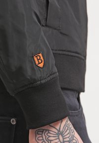 Svart bomberjacka med ribbad stickad mudd och en orange broderad logotyp på ärmen. Slät, lättviktsmaterial med minimal textur.