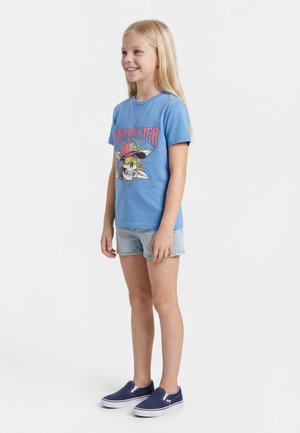 Jeune fille souriante, debout, portant un t-shirt graphique bleu avec un crâne et du texte, un short en jean clair et des baskets slips-on bleu marine.