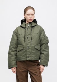 Olišu zaļa izguldīta jaka ar rāvējslēdzēja kapuci, priekšējiem pogām, divām kabatām uz krūtīm un aproces nodalījumu kreisajā piedurknē.