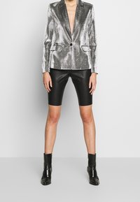 Blazer metálico plateado con solapas de muesca y dos bolsillos frontales, combinado con pantalones cortos de cuero negros y botas ankle.