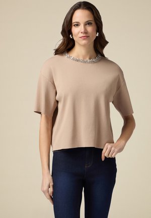 Beiger, kurzer Pullover aus Strickstoff mit einem runden Halsausschnitt, der mit klaren Strasssteinen verziert ist, kombiniert mit dunkelblauen Jeans.