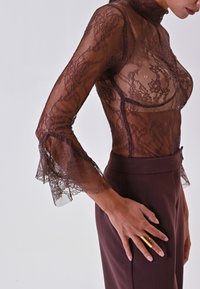 Top lungo in pizzo marrone con maniche a kimono, collo alto e polsini svasati, abbinato a pantaloni marroni a vita alta e un anello d'oro sulla mano destra.