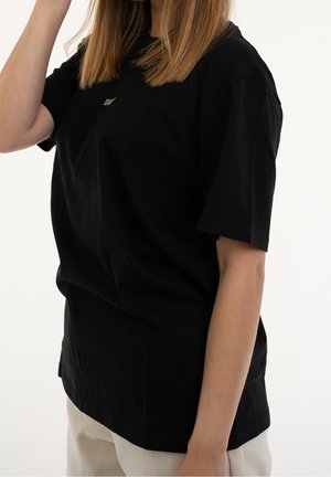 Femme aux cheveux lisses châtain clair portant un T-shirt noir ample et un pantalon de couleur claire sur un fond blanc uni.