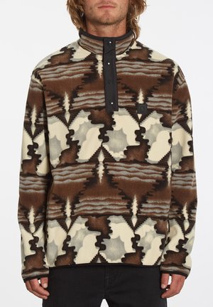 Mann trägt einen braun-creme-grau gemusterten Fleecepullover mit schwarzer Druckknopfleiste und Stehkragen, steht vor weißem Hintergrund.