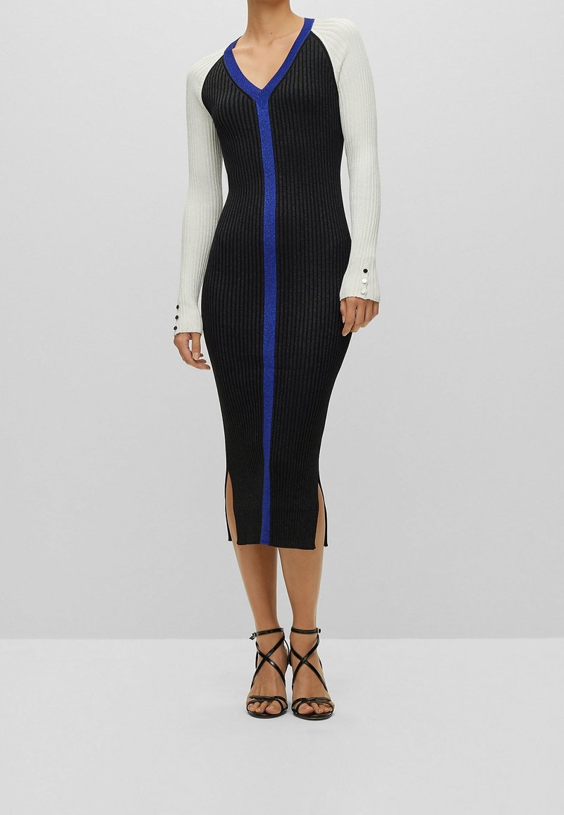 Femme portant une robe midi noire côtelée avec des manches longues blanches, une bordure bleue sur l’avant, des fentes latérales et des sandales à talons hauts noires à brides.