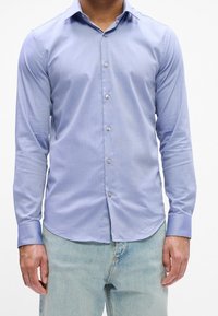 Homme portant une chemise bleu clair ajustée à manches longues et un jean bleu clair, debout les bras détendus le long du corps.