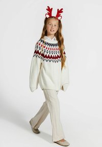 Dziewczyna idzie, ubrana w biały sweter z wzorem, kremowe spodnie w warkocze, beżowe buty oraz czerwony brokatowy opaska na głowę z rogami renifera.