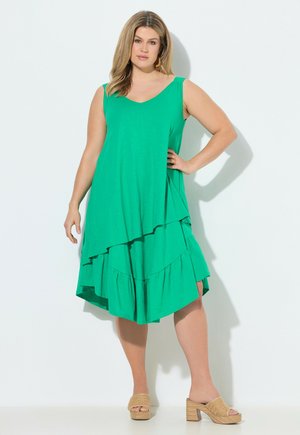 Kvinde med langt hår iført en ærmeløs, grøn lagdelt kjole og beige platformsandaler, stående med den ene hånd på hoften.