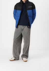 Veste gonflante avec une partie supérieure noire et une partie inférieure bleue, associée à un pantalon gris à rayures larges et des baskets crème avec des rayures noires.