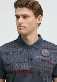 Gray polo shirt with a textured fabric, featuring embroidered graphics in various colors, buttons on the collar, and a patch logo on the chest.