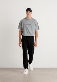 Grå bomulds t-shirt med sort "Calvin Klein" logo, parret med sorte joggers med elastisk talje og hvide sneakers, båret af en stående model.