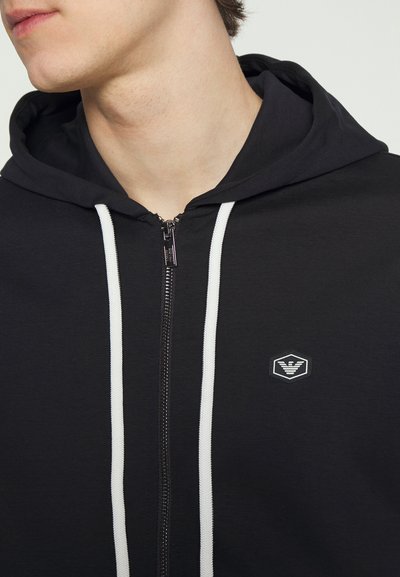 Black zip-up hoodie with white drawstrings, featuring a hexagonal logo on the chest, made from a smooth, textured fabric.
