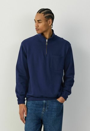 Junger Mann mit geflochtenem Haar, der einen marineblauen Pullover mit Reißverschluss am Kragen und blaue Jeans trägt, steht mit einer Hand in der Tasche und schaut nach links.