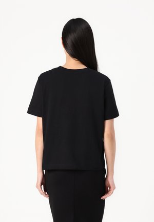 Personne aux longs cheveux noirs portant un t-shirt noir uni à manches courtes et une jupe noire, de dos sur un fond blanc.