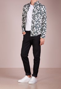 Floral patterned shirt with green and white shades, paired with black tapered jeans and white sneakers. Model stands with hands in pockets.