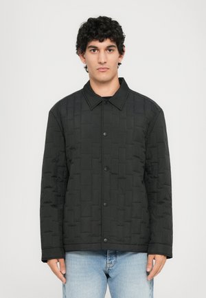 Black quilted jacket with a collared neckline, featuring a button closure and textured geometric pattern, paired with blue denim jeans.