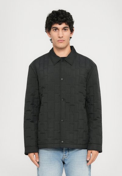 Veste matelassée noire avec un col, dotée d'une fermeture à bouton et d'un motif géométrique texturé, associée à un jean en denim bleu.