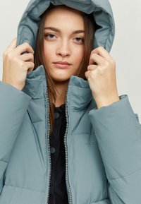 Padded jacket in light blue with a high collar, zip closure, and hood. Features a textured fabric and decorative buttons.