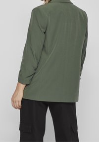 Olivově zelený blazer se strukturovaným designem, dlouhými rukávy a jedním rozventilováním na zadní straně, zkombinovaný s volnými černými cargo kalhotami.