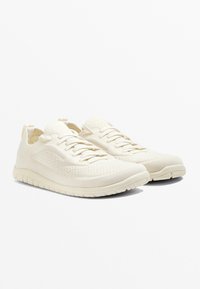 Ljusbeige sportskor i meshmaterial, med flexibel sula, snörning och dragflikar på hälen för enkel på- och avtagning.