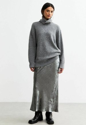 Pull en tricot gris avec col montant, associé à une jupe argentée métallisée longue jusqu'aux chevilles. Des bottines noires à la cheville complètent la tenue.