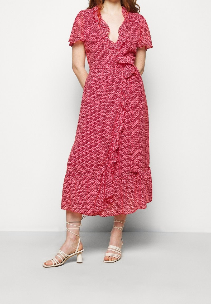 Femme portant une robe portefeuille rouge avec de petits motifs blancs, des détails à volants, des manches courtes et des sandales à talons beige avec des brides à la cheville.