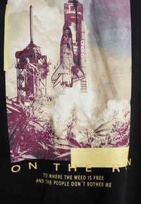 Svart t-shirt med en grafisk bild av en rymdraketuppskjutning med ett torn, omgiven av moln. Text inkluderar "PÅ R" och en fras om frihet.