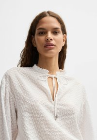 Blusa bianca strutturata con colletto a volants e scollatura a occhio di bue, dotata di un cordoncino che termina con piccole punte metalliche.