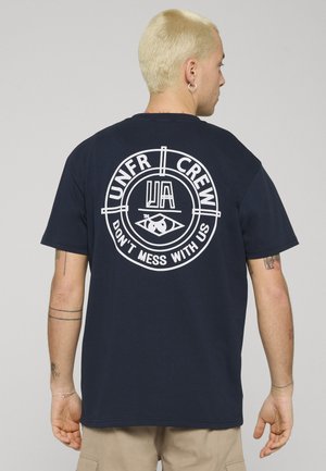 Person mit kurzen blonden Haaren, die ein marineblaues T-Shirt mit der Aufschrift „UNFR CREW“ und „DON'T MESS WITH US“ in einem kreisförmigen Design auf dem Rücken trägt.