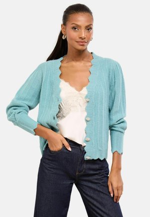 Light blue knitted cardigan with scalloped edges, button front, and ribbed texture, layered over a white lace-trimmed top and dark jeans.