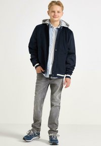 Marineblaues Jackett mit grauem Kapuzenpullover, knöpfen an der Vorderseite, weißen gestreiften Manschetten; hellblaues gestreiftes Hemd darunter; graue Jeans und blaue Sneaker.