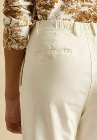 Pantaloni de culoare crem, cu talie elastică, având un singur buzunar la spate cu nasture și o textură netedă. Cusături laterale vizibile.