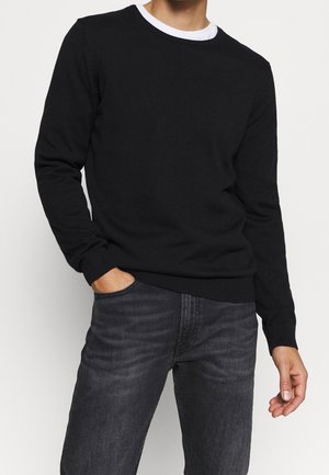 Mann trägt schwarzen Langarmpullover über weißem Hemd und dunkelgrauen Jeans, eine Hand in der Tasche, die andere entspannt an der Seite.