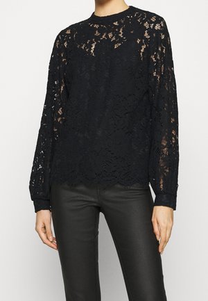 Kobieta ubrana w czarną koronkową bluzkę w kwiatowy wzór z długim rękawem, połączoną z dopasowanymi czarnymi skórzanymi spodniami, stojąca na jednolitym tle.