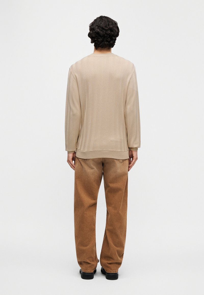 Haut beige texturé à manches longues avec un col rond, associé à un pantalon en velours côtelé marron ample. La tenue présente un motif chevrons subtil.