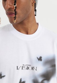 Camiseta blanca en mezcla de algodón con escote redondo. Presenta un estampado gráfico difuminado y el texto "VISIÓN DIFUSA" en letras negras en negrita.