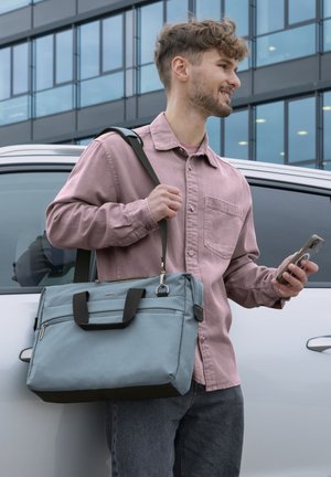 Junger Mann im pinken Hemd hält Smartphone und graue Umhängetasche, steht neben weißem Auto und modernem Glasgebäude.