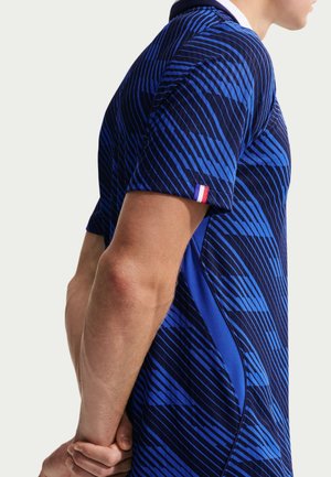 Homme portant une chemise à manches courtes bleue à motifs avec une petite étiquette du drapeau français sur la manche, les mains jointes dans le dos.