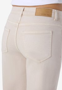 Μπεζ τζιν ψηλόμεσα με πίσω τσέπες και καφέ δερμάτινη ετικέτα με την επιγραφή "NOISY MAY", φορεμένα από ένα άτομο με λευκό τοπ.