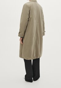 Langer, khakifarbener Mantel aus leichtem Stoff, mit gerafftem Saum, geknöpften Manschetten und einem Rückenschlitz. Kombiniert mit schwarzen, weiten Hosen.