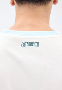 Achteraanzicht van een persoon die een wit gestructureerd shirt draagt met een lichtblauwe kraag en "ÖSTERREICH" in blauwgroen op de bovenrug gedrukt.