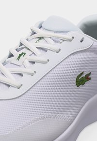 Λευκό αθλητικό παπούτσι με πράσινο κεντητό λογότυπο κροκόδειλου στο πλάι και στη γλώσσα, με λευκά κορδόνια και υφή από πλεκτό δίχτυ.