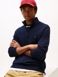Hanorac închisoare cu fermoar, de culoare bleumarin, cu manșete din tricot, asortat cu un tricou alb. Șapcă roșie cu logo pe față. Modelul are brațele încrucișate, pe un fundal deschis.