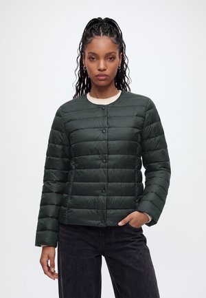 Femme aux cheveux tressés portant une doudoune verte foncée et un pantalon noir, debout avec une main dans la poche sur fond blanc.