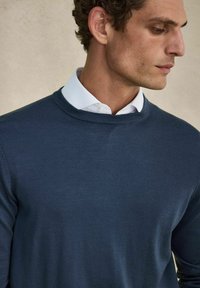 Mörkblå stickad tröja med rund halsringning, bärs över en vit skjorta med krage. Slät textur, kroppsnära design som visar en modern avslappnad stil.