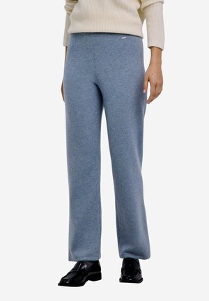 Persoon draagt lichtblauwe, wijd uitlopende gebreide broek, crème geribde trui en zwarte leren loafers, staand tegen een witte achtergrond.